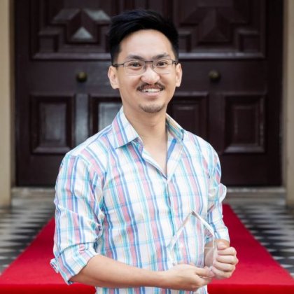 A man with black spiky hair and glasses smiles broadly outside double doors with a red carpet behind him. He is holding a glass trophy.